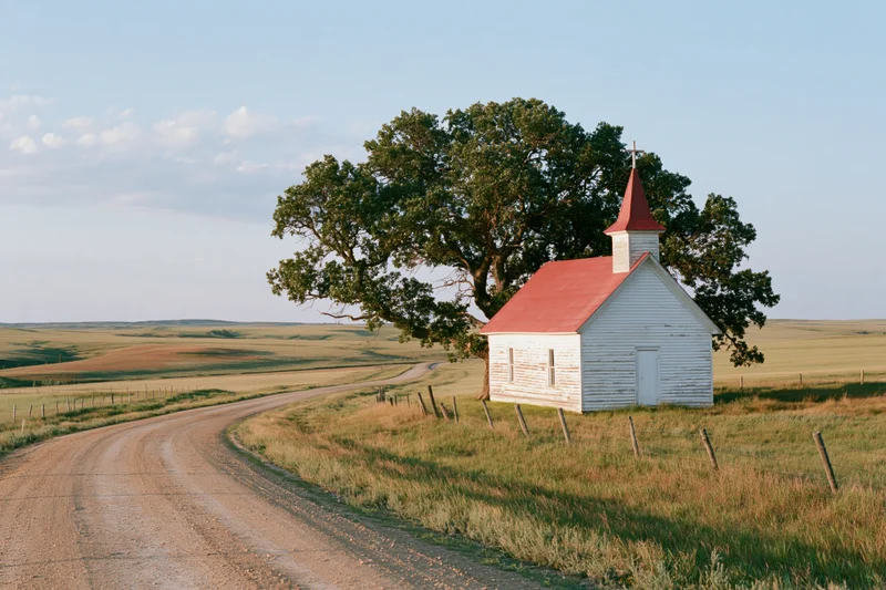 Small white country church with red roof and steeple beside a winding dirt road in a grassy field