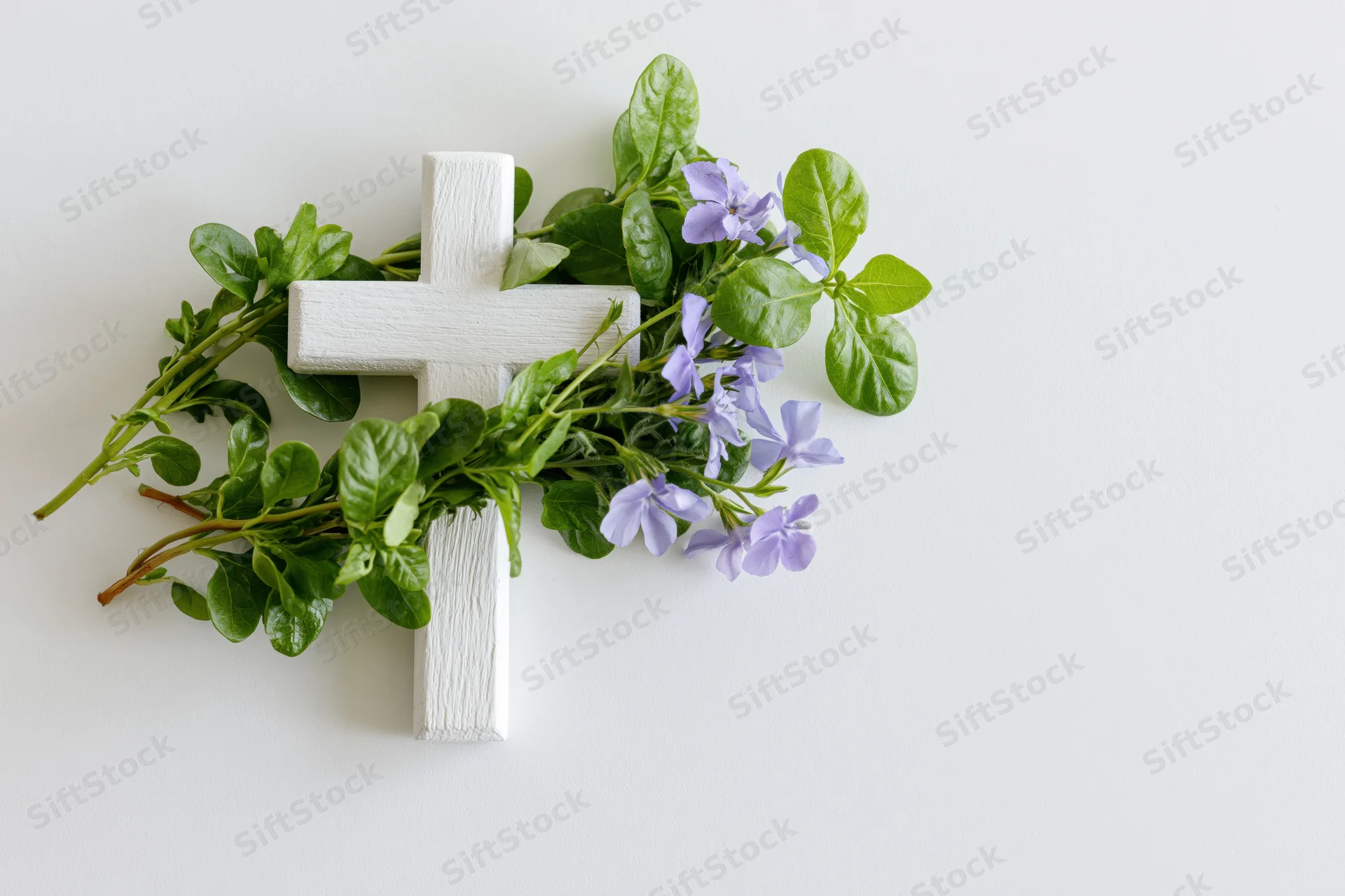 White wooden cross adorned with green leaves and purple flowers on a plain white surface