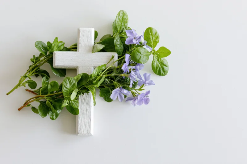White wooden cross adorned with green leaves and purple flowers on a plain white surface