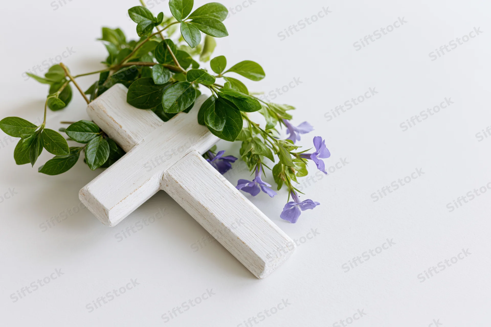 White wooden cross with green leaves and purple flowers arranged on a white surface