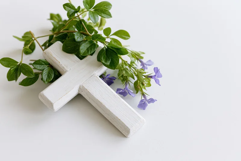White wooden cross with green leaves and purple flowers arranged on a white surface