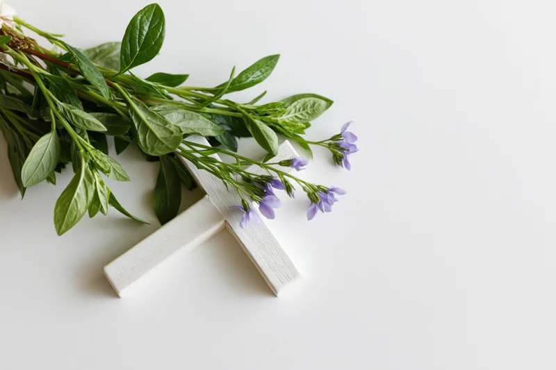 Small white painted wooden cross lying flat on white surface with purple flowers and green leaves arranged beside it