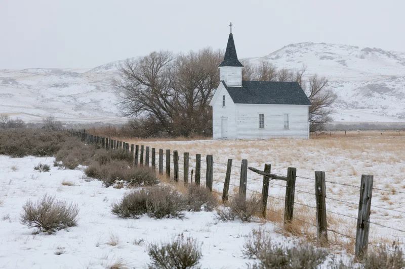 Small white rural church with dark steeple and cross standing in snowy field behind wooden fence with snow-covered mountains in background