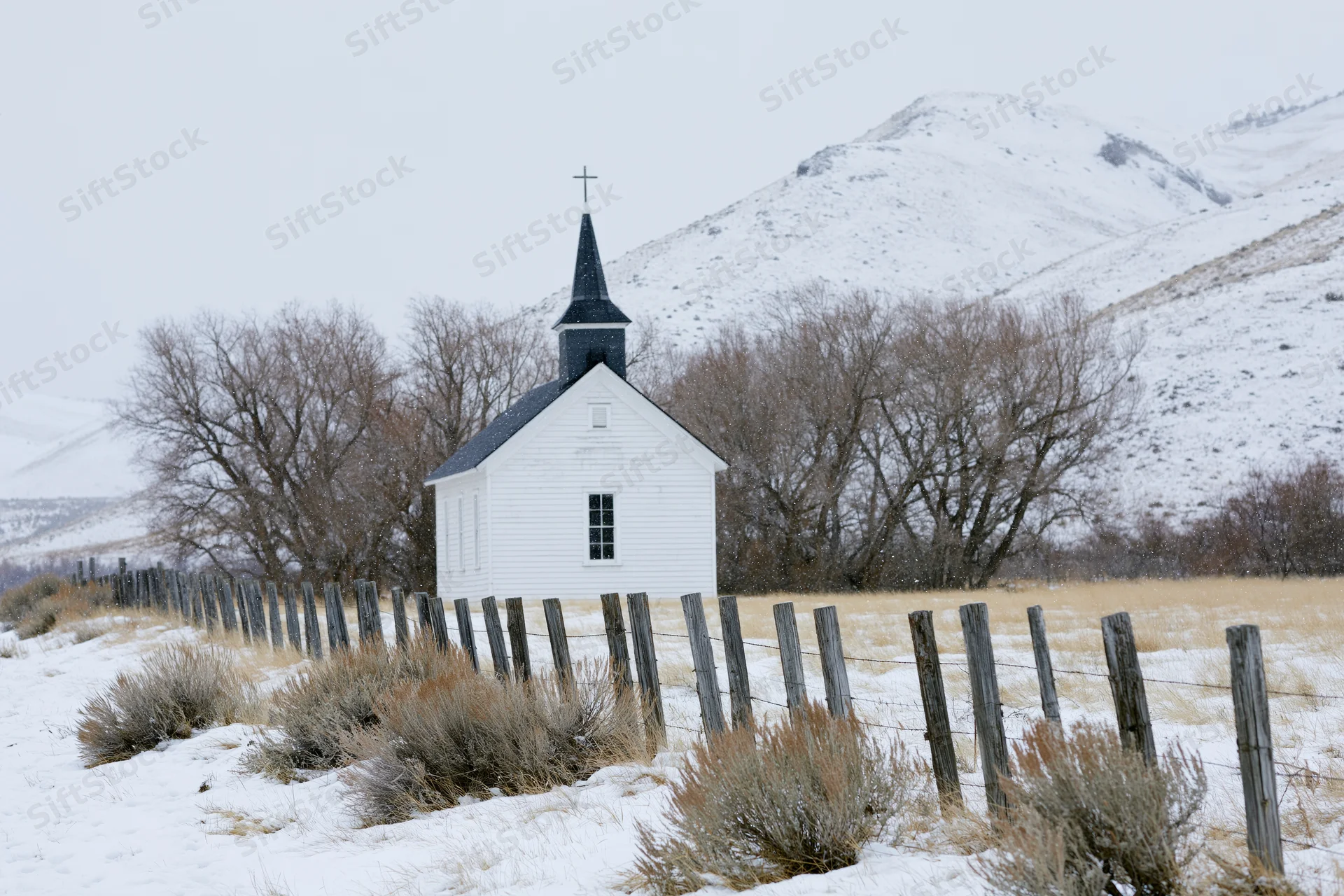 Small white rural church with dark steeple and cross standing in snowy field near mountains