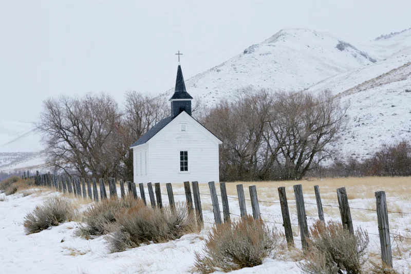 Small white rural church with dark steeple and cross standing in snowy field near mountains