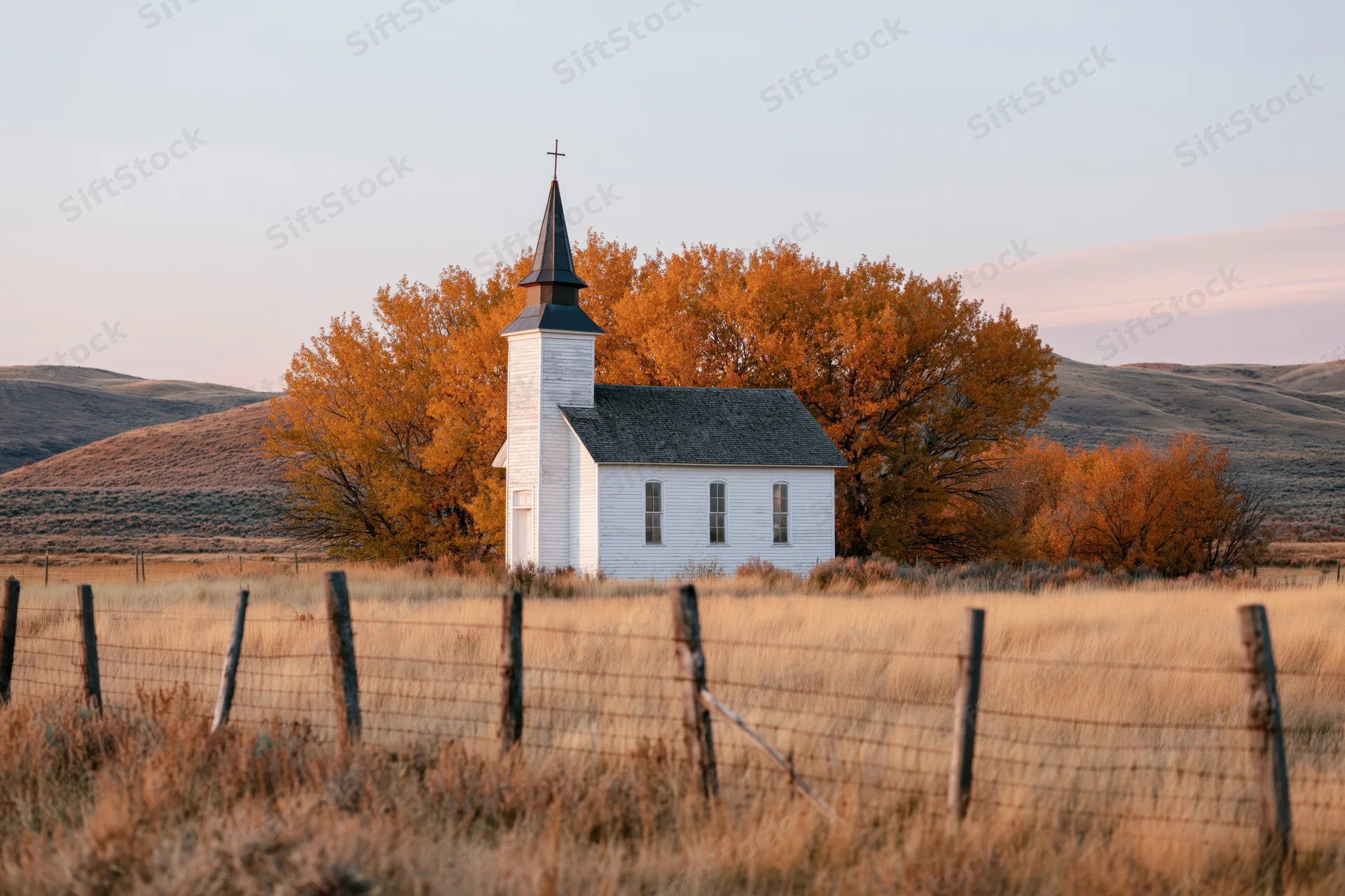 Small white rural church with dark steeple and cross standing in a fenced field surrounded by autumn trees and rolling hills