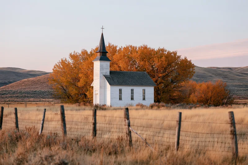 Small white rural church with dark steeple and cross standing in a fenced field surrounded by autumn trees and rolling hills