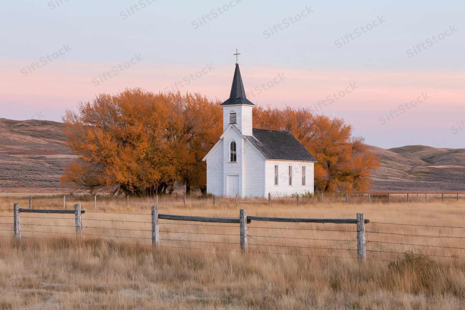 Small white rural church with dark steeple and cross standing in a fenced field surrounded by autumn trees and rolling hills at sunset
