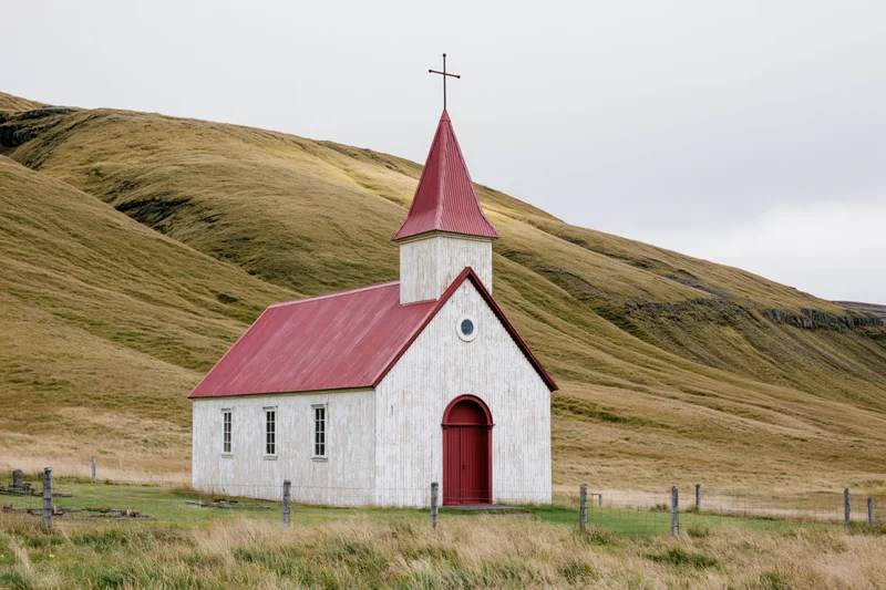 Small white church with red corrugated metal roof and steeple in a grassy rural landscape with rolling hills
