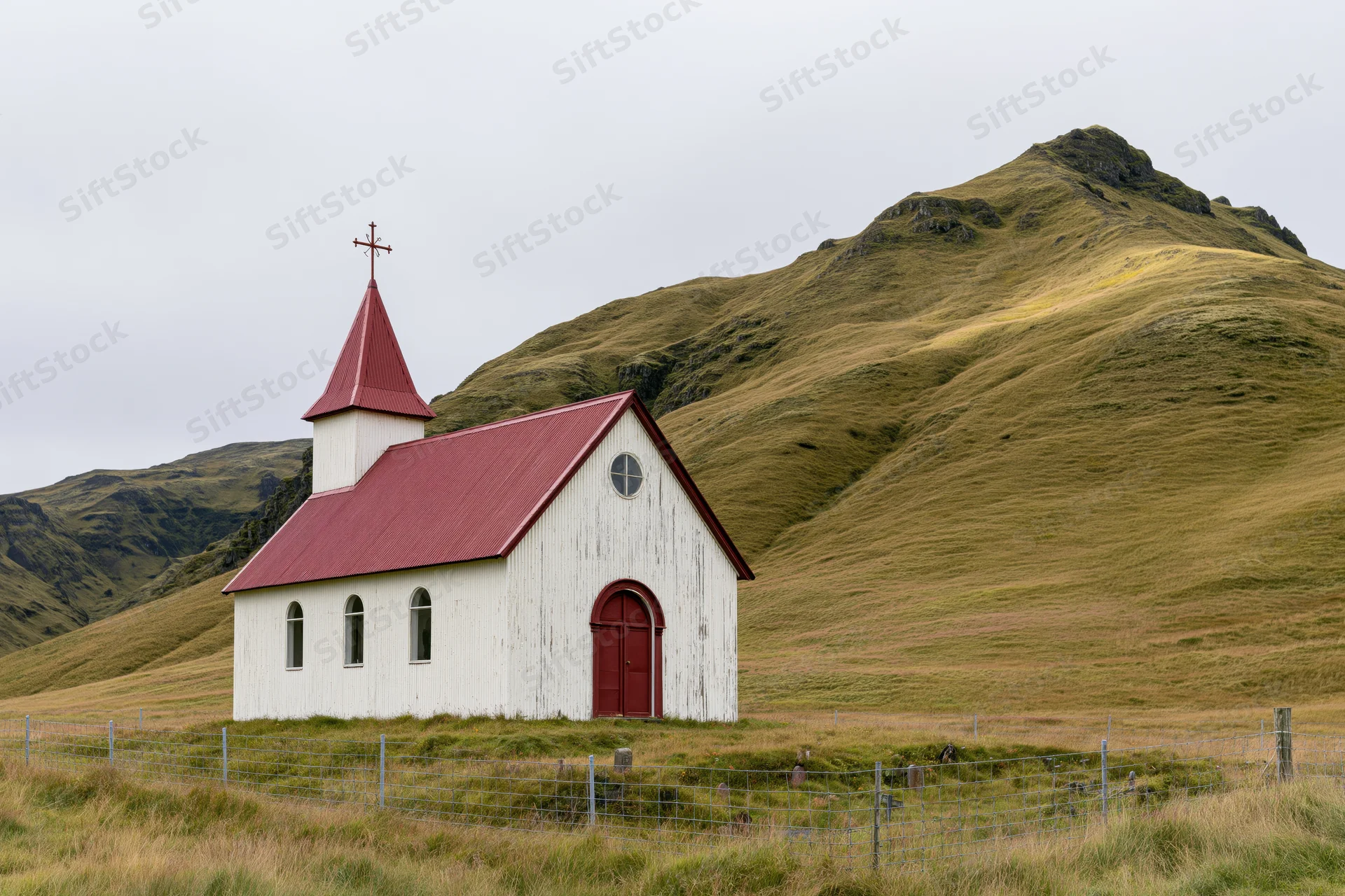 Small white rural church with red corrugated metal roof and steeple set against grassy hills and a fenced graveyard