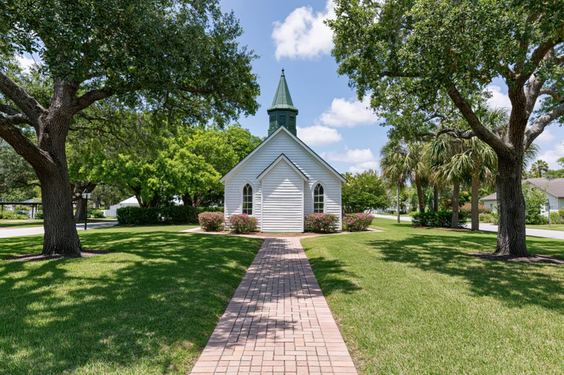 Small white wooden chapel with green steeple centered along brick pathway through grassy lawn lined with trees
