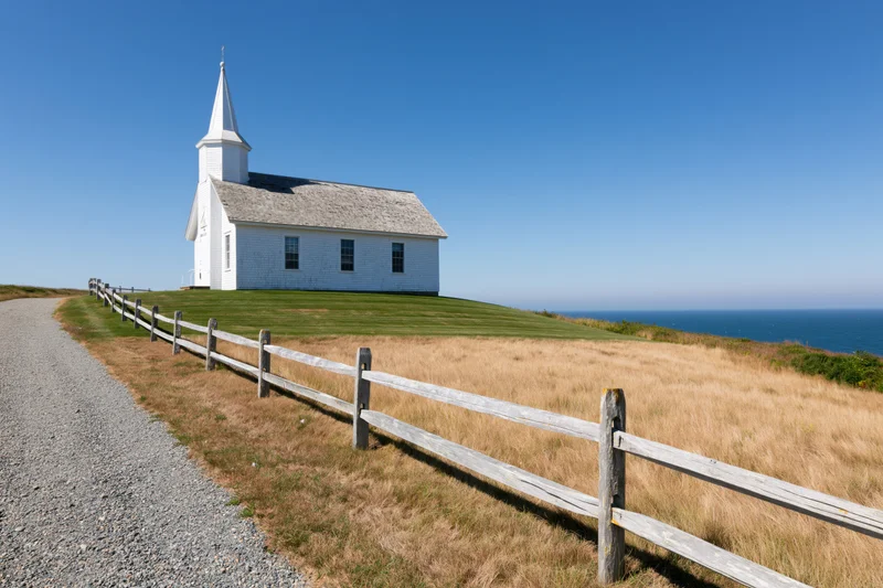 White wooden church with pointed steeple on grassy hill overlooking ocean, beside gravel path and wooden fence