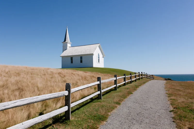 Small white wooden church with pointed steeple on grassy hill overlooking ocean, accessed by gravel path beside wooden fence