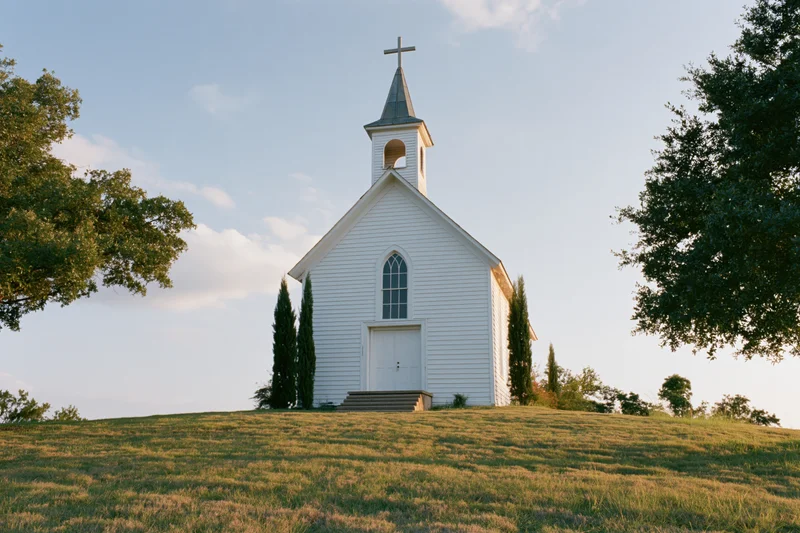 Small white wooden church with steeple and cross on grassy hill surrounded by trees under blue sky
