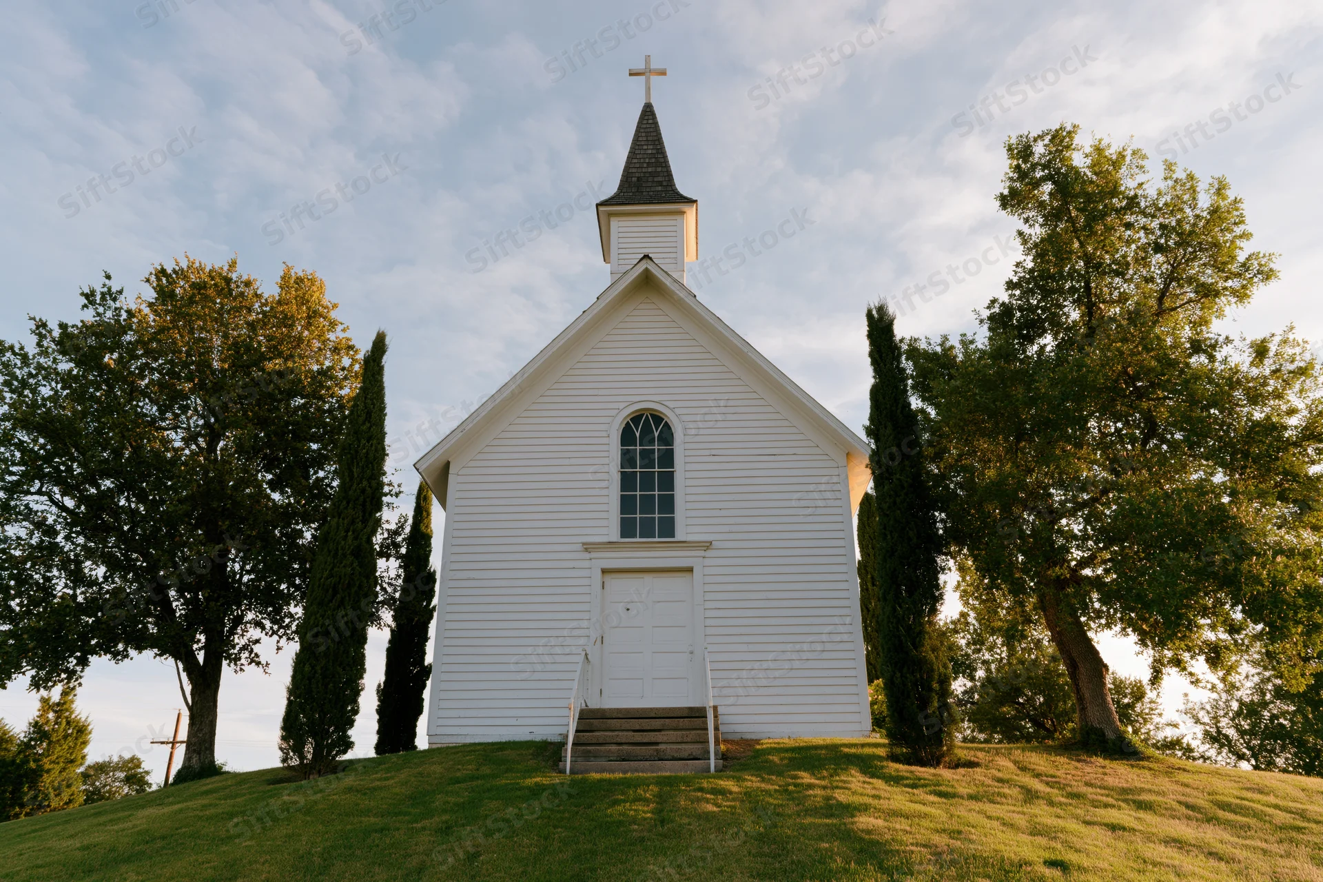 Small white wooden church with steeple and cross on grassy hill surrounded by trees