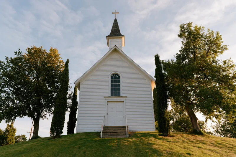 Small white wooden church with steeple and cross on grassy hill surrounded by trees