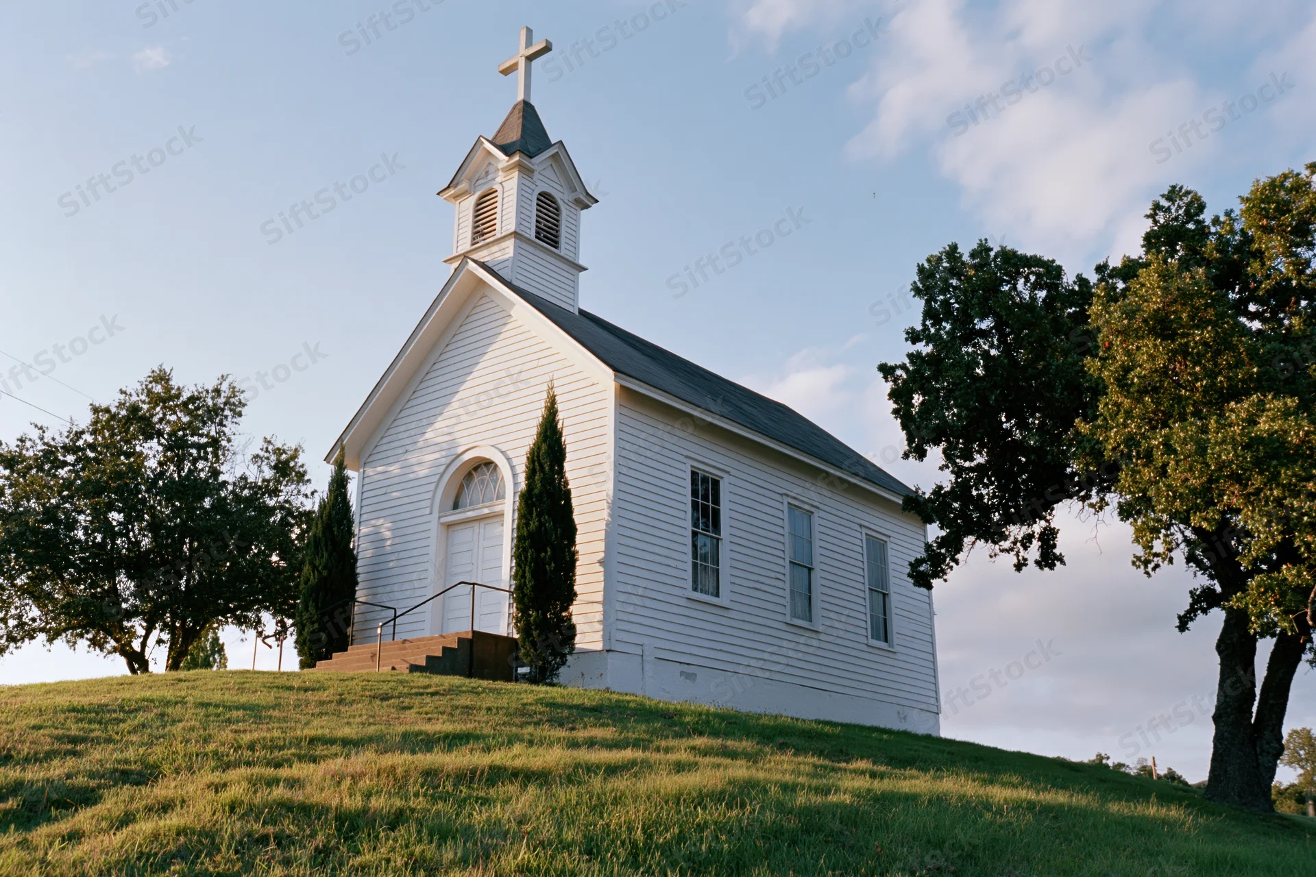 Small white wooden church with steeple and cross on grassy hill surrounded by trees under blue sky