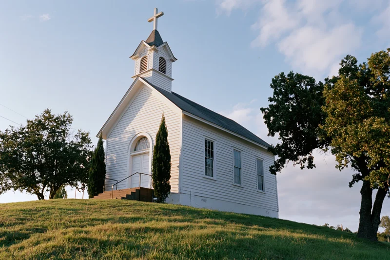 Small white wooden church with steeple and cross on grassy hill surrounded by trees under blue sky
