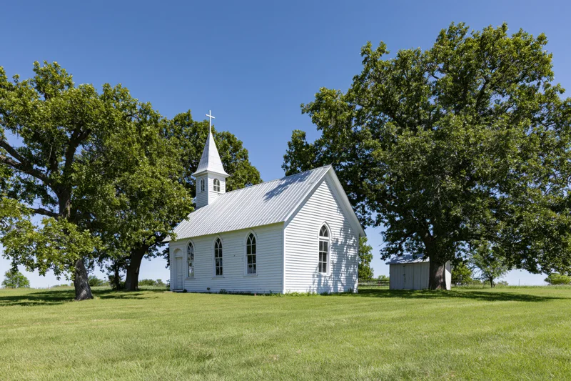 Small white wooden church with steeple and cross in a grassy rural field surrounded by trees under a clear blue sky