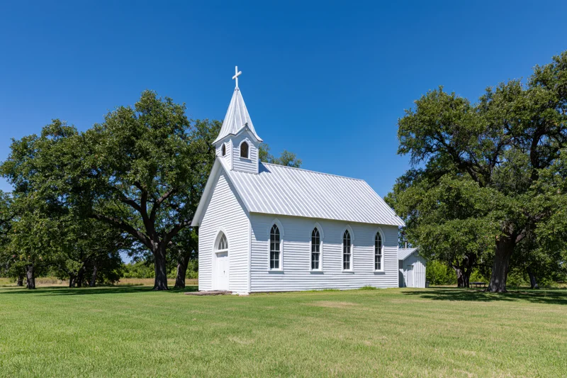 Small white wooden church with steeple and cross in a grassy field surrounded by trees under a clear blue sky