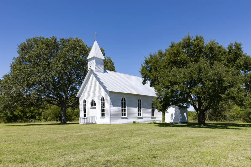 Small white wooden church with steeple and cross in a grassy rural field surrounded by trees under a clear blue sky