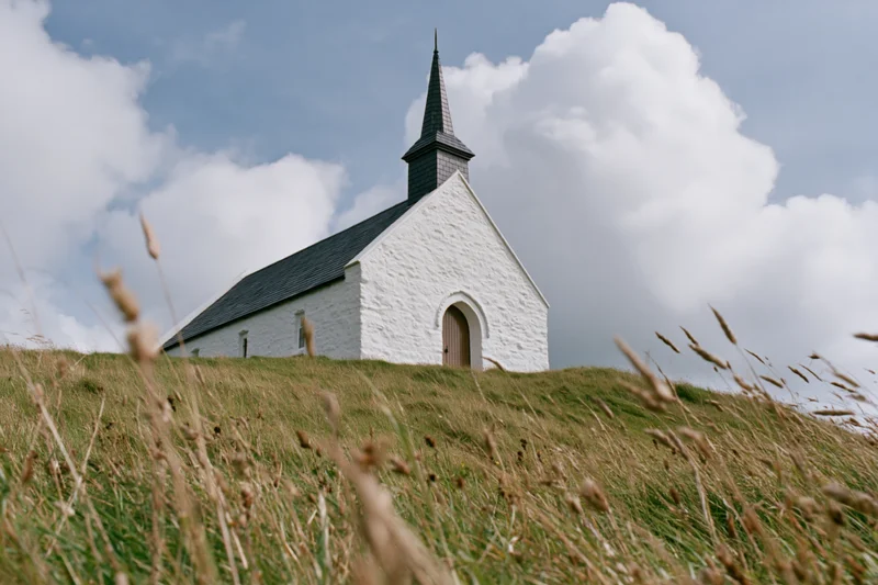 Small whitewashed chapel with dark pointed steeple on a grassy hill under cloudy sky