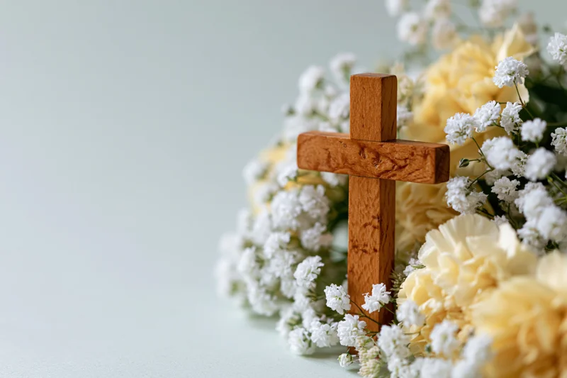 Wooden cross standing among white and yellow flowers on a light surface