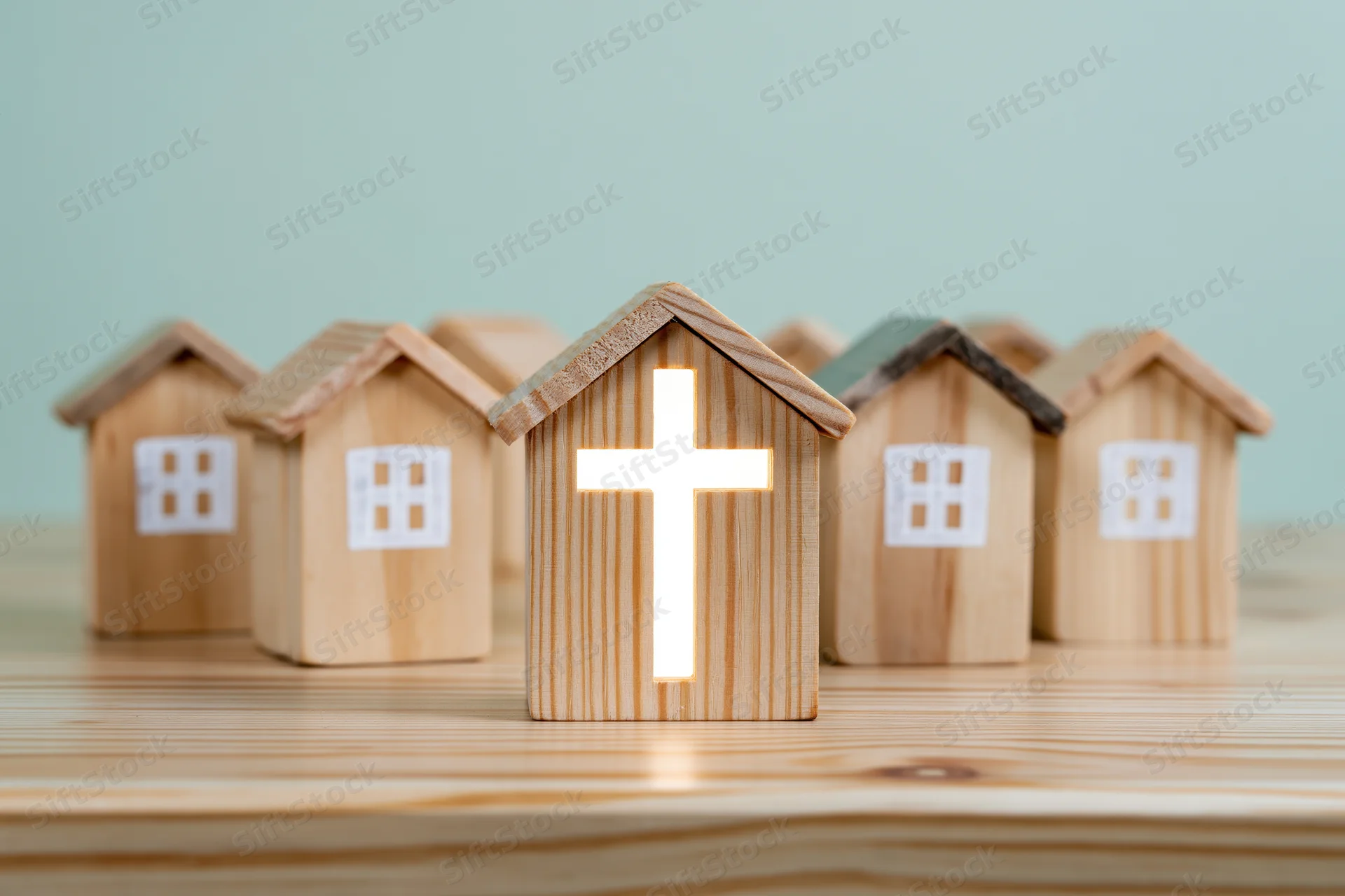 Wooden house miniatures in a row with a glowing cross on the front one on a light wooden surface