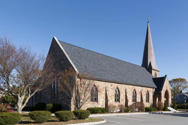 Stone church with steeple and cross under clear blue sky in suburban setting