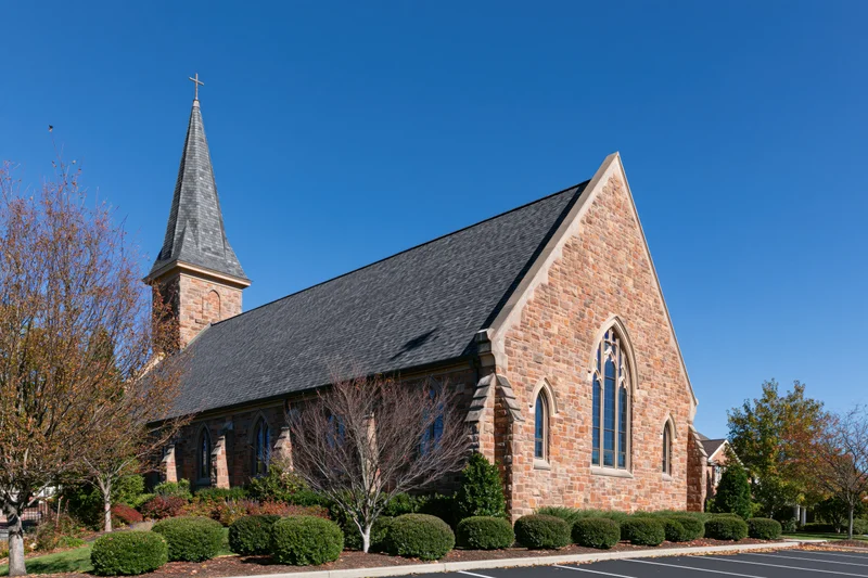 Stone church with steeple and cross under clear blue sky in suburban setting