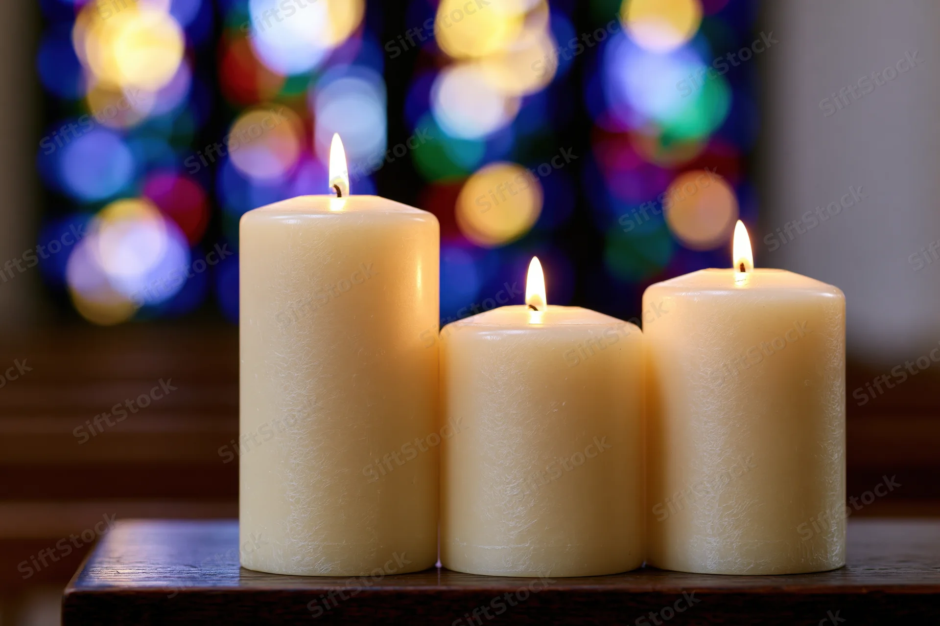 Three cream pillar candles burning with warm flames on a wooden table in front of a stained glass window