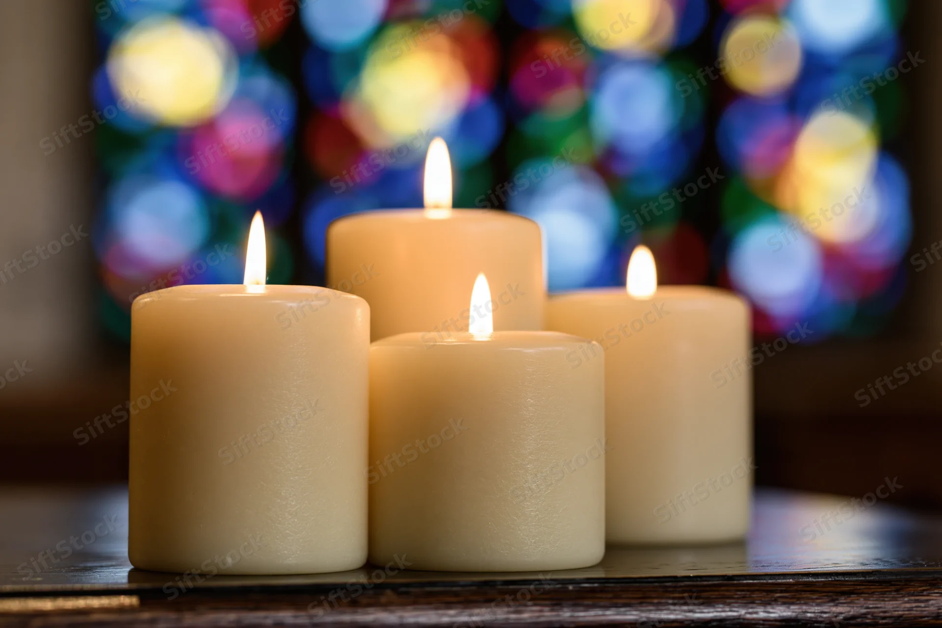 Four cream pillar candles burning with warm flames on a wooden table in front of a stained glass window