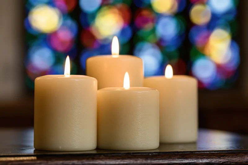 Four cream pillar candles burning with warm flames on a wooden table in front of a stained glass window