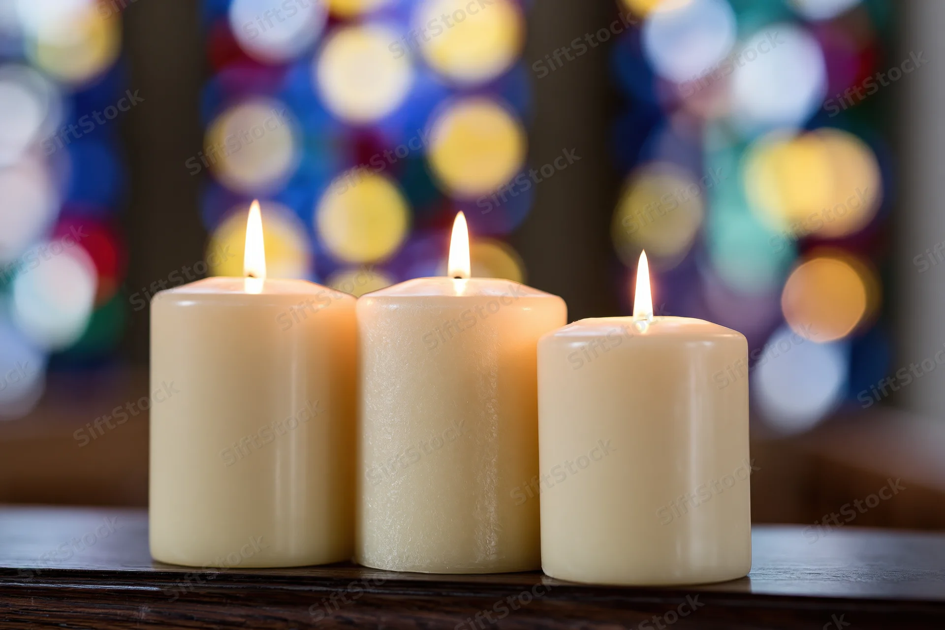 Three cream pillar candles burning with warm flames on a wooden table in front of a stained glass window