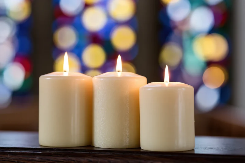 Three cream pillar candles burning with warm flames on a wooden table in front of a stained glass window