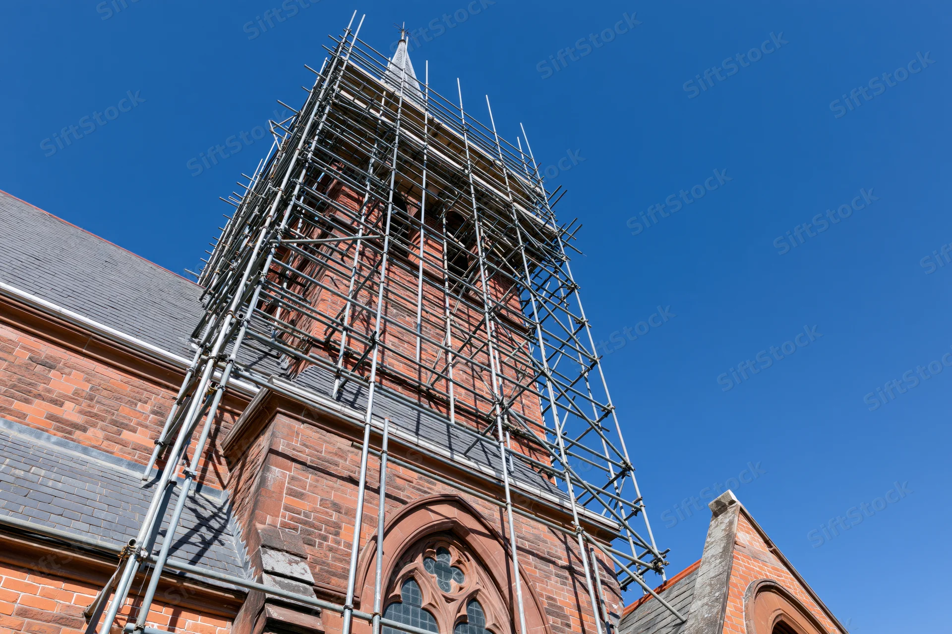Victorian red-brick church tower under restoration with steel scaffolding against clear blue sky