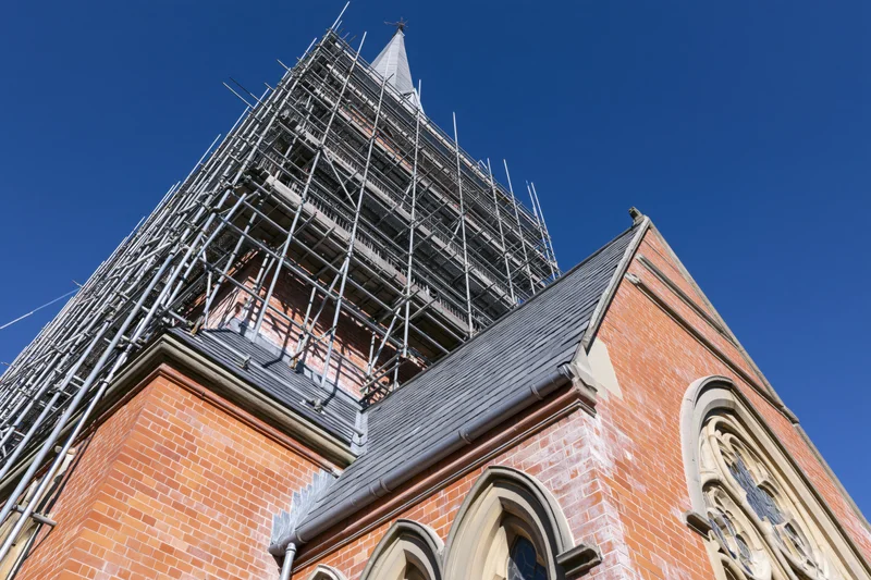 Victorian red-brick church under restoration with steel scaffolding on its tower against a clear blue sky