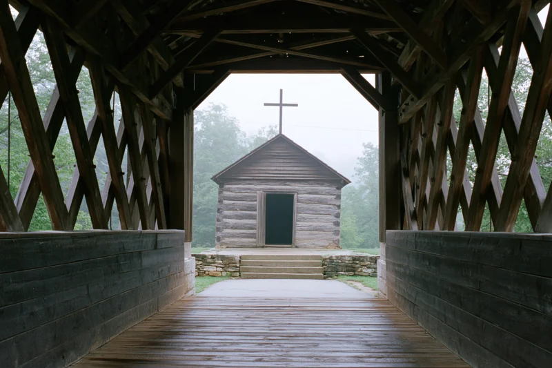 View from inside wooden covered bridge looking out at small log church with cross in misty forest