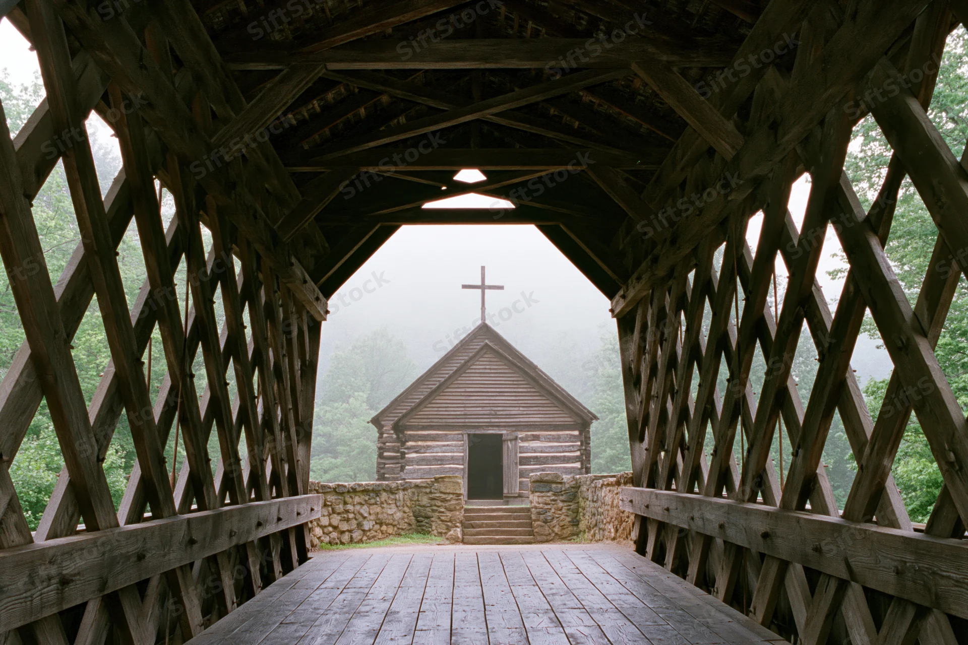 View from inside wooden covered bridge looking toward foggy log church with cross on roof