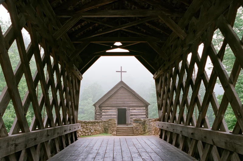 View from inside wooden covered bridge looking toward foggy log church with cross on roof