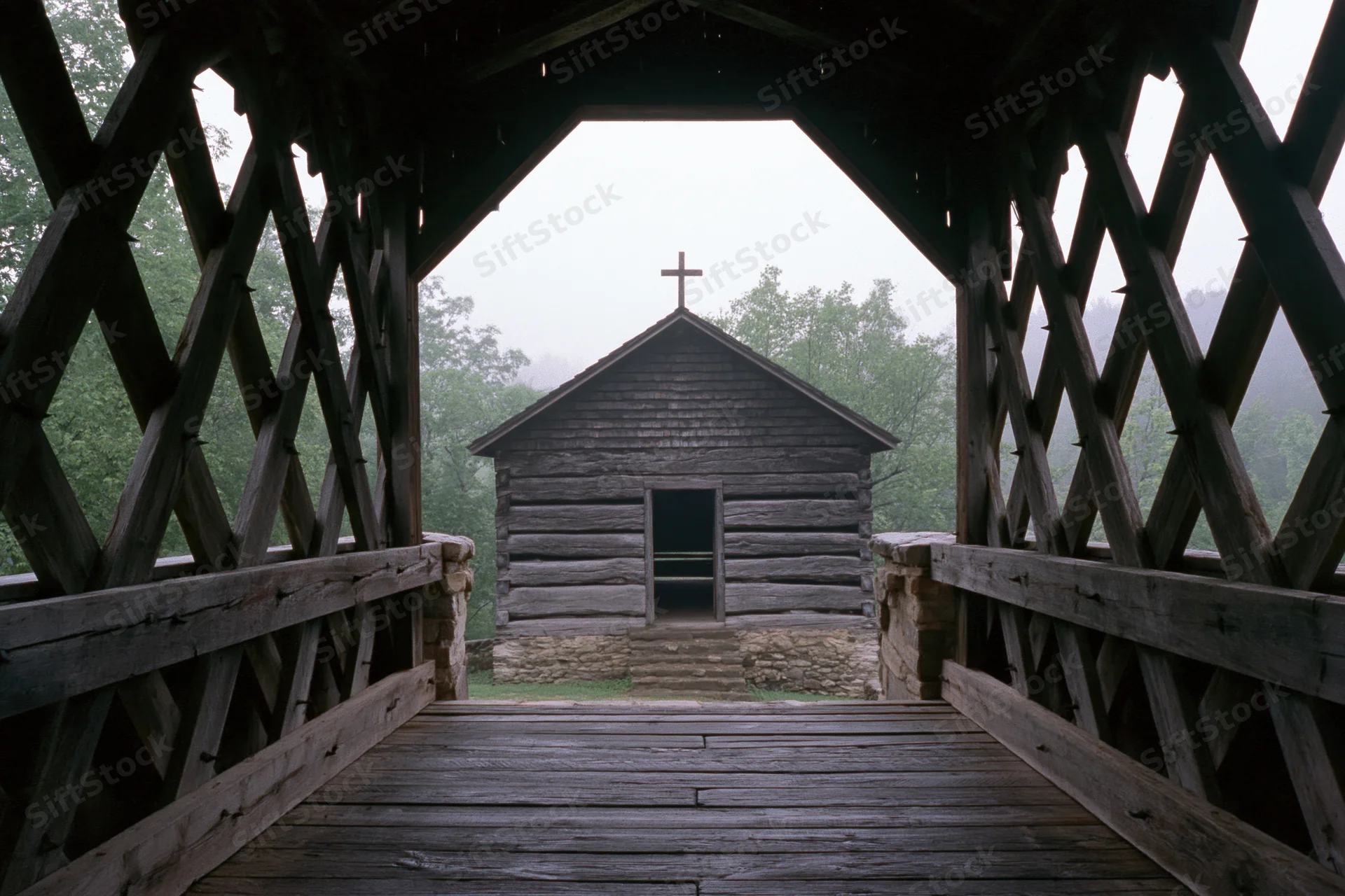 View from inside wooden covered bridge looking toward small log church with cross on roof in misty forest setting