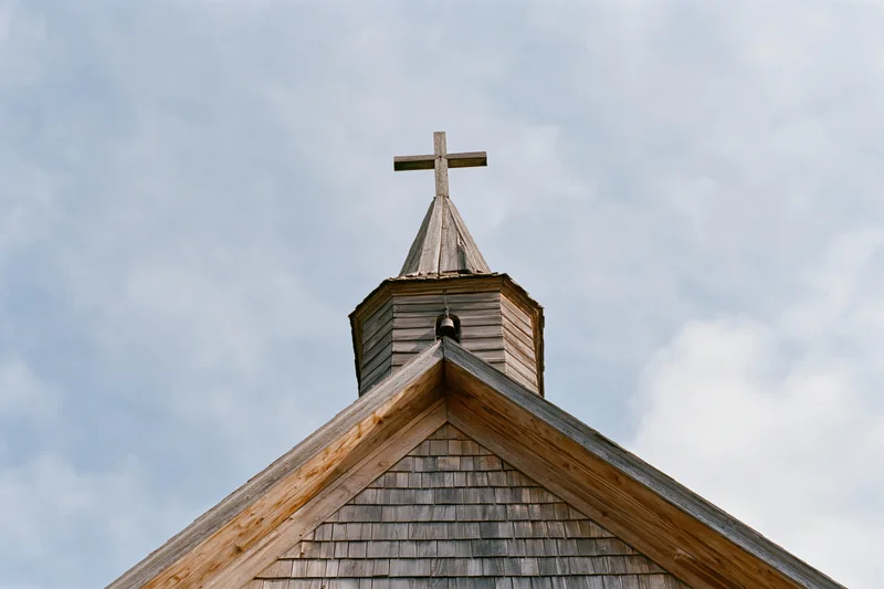 Weathered wooden church steeple with a cross at its peak against a cloudy sky