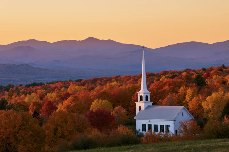 White steepled church surrounded by colorful autumn trees with mountains in the background at sunset