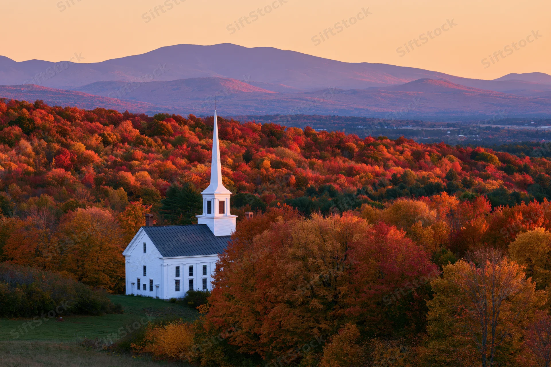 White steepled church surrounded by colorful autumn trees in a rural landscape with mountains at sunset