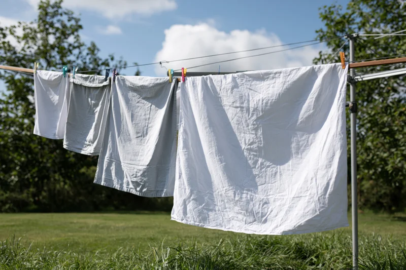White and light grey laundry drying on a freestanding clothesline in a grassy yard under a blue sky with clouds