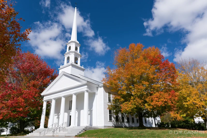 White clapboard church with tall steeple and columns surrounded by autumn trees under blue sky
