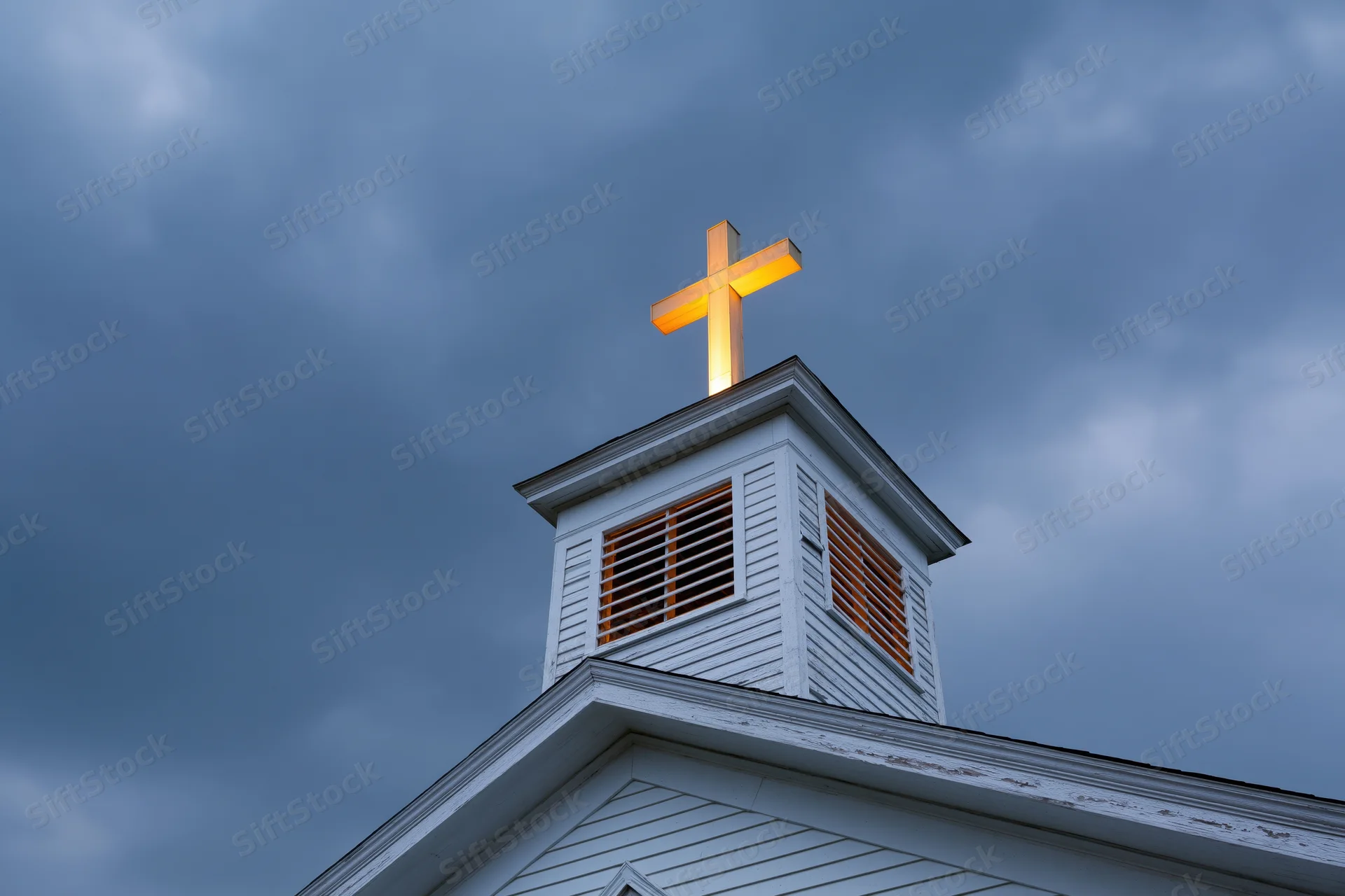 Illuminated cross atop white wooden church steeple against cloudy sky at dusk