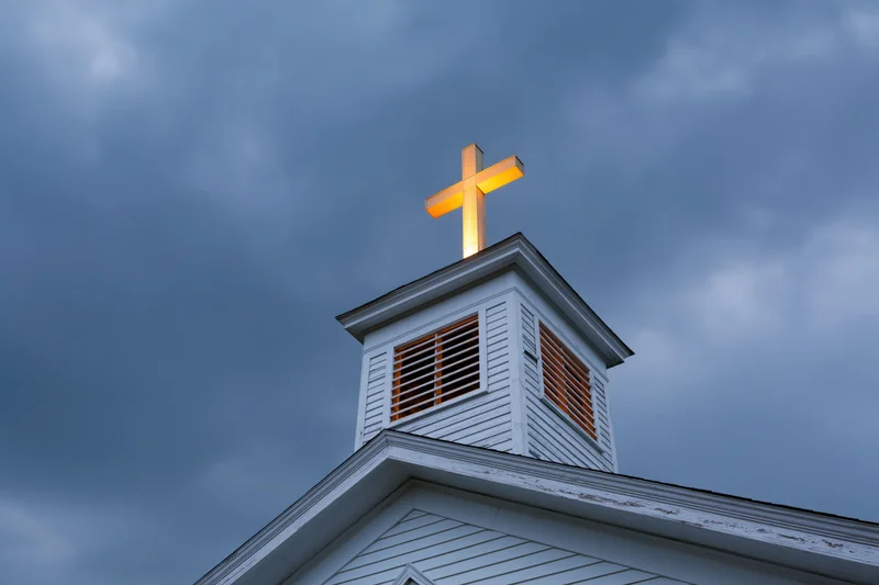 Illuminated cross atop white wooden church steeple against cloudy sky at dusk