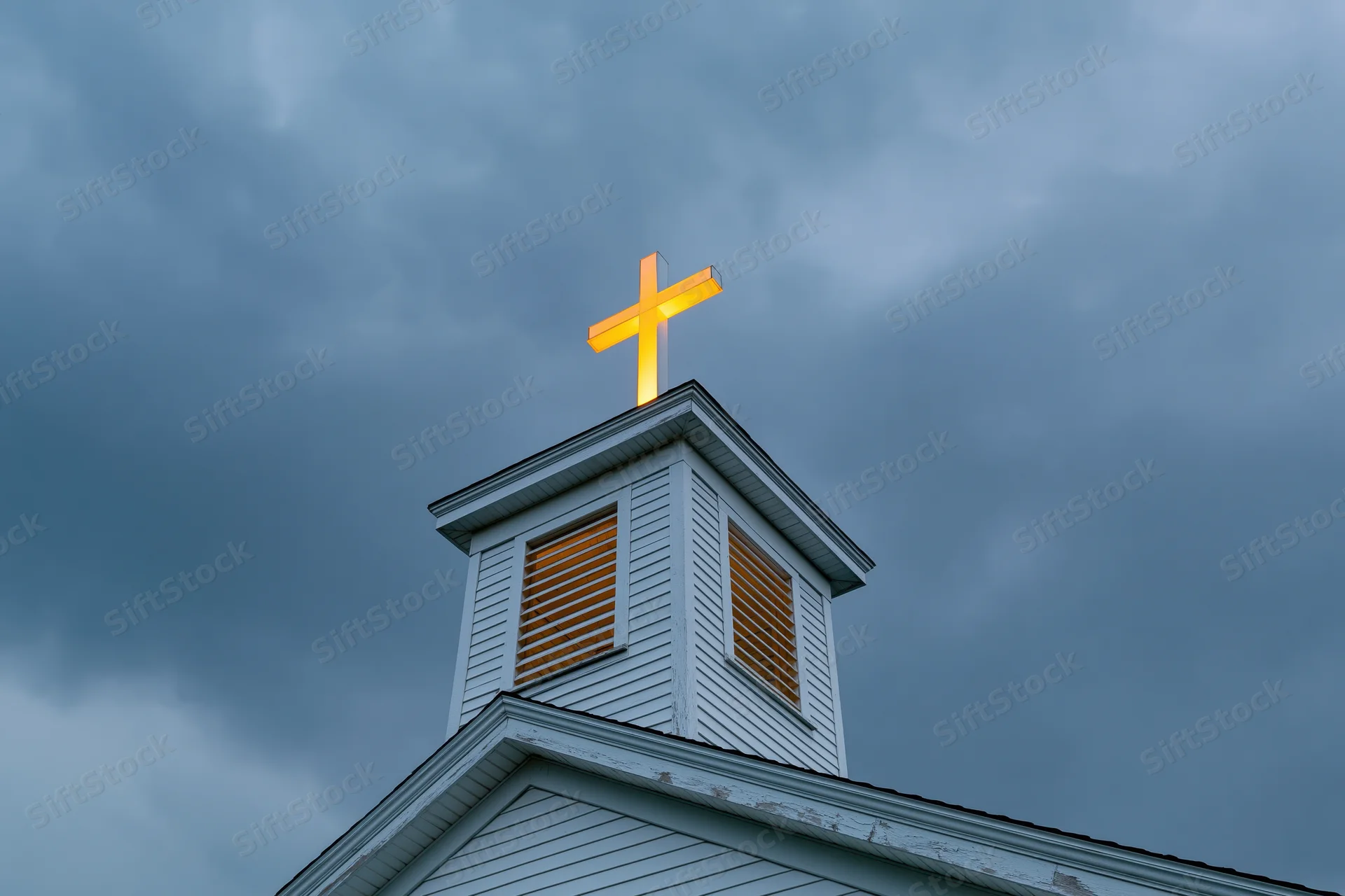 Illuminated cross atop white wooden church steeple against cloudy sky