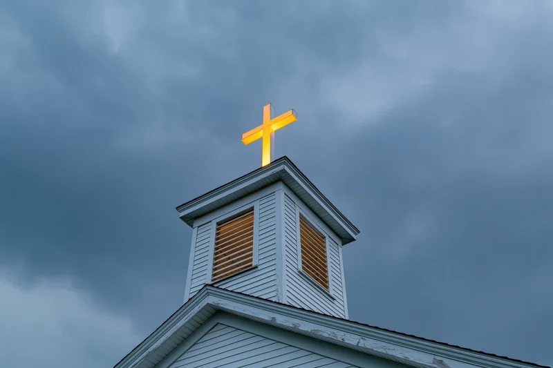 Illuminated cross atop white wooden church steeple against cloudy sky